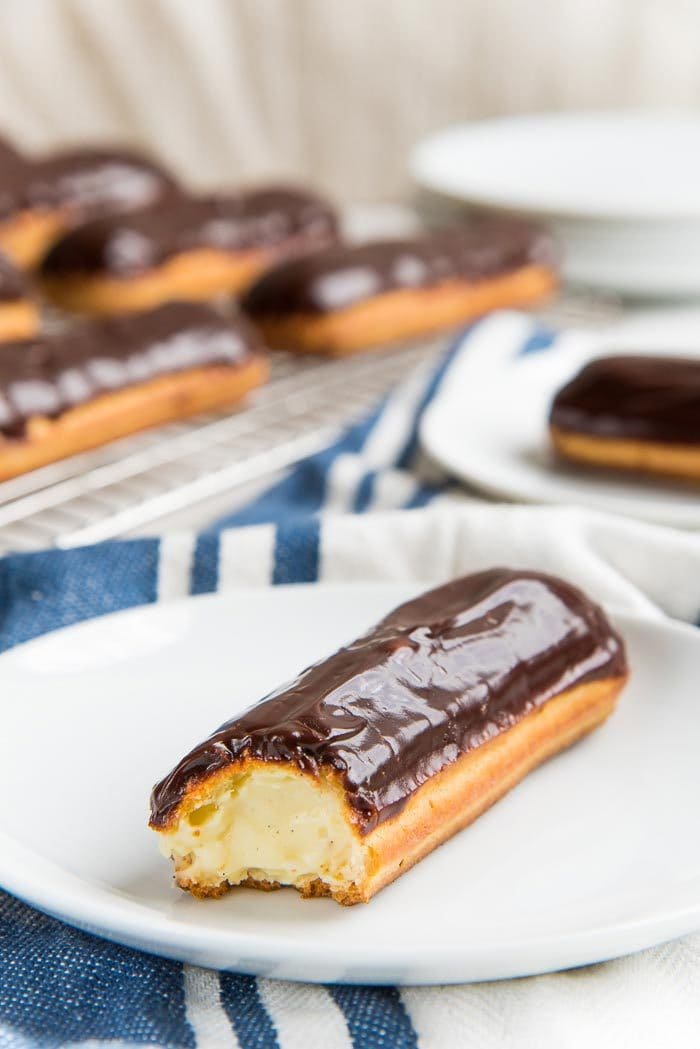 A close up of a chocolate eclair with vanilla pastry cream filling, with a bite taken out, placed on a white plate.