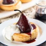 Chocolate stuffed poached pear tart served on a white plate with mascarpone cream and red wine sauce.