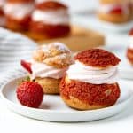 Two strawberry cream puffs on a plate, one with a red craquelin top and one without.