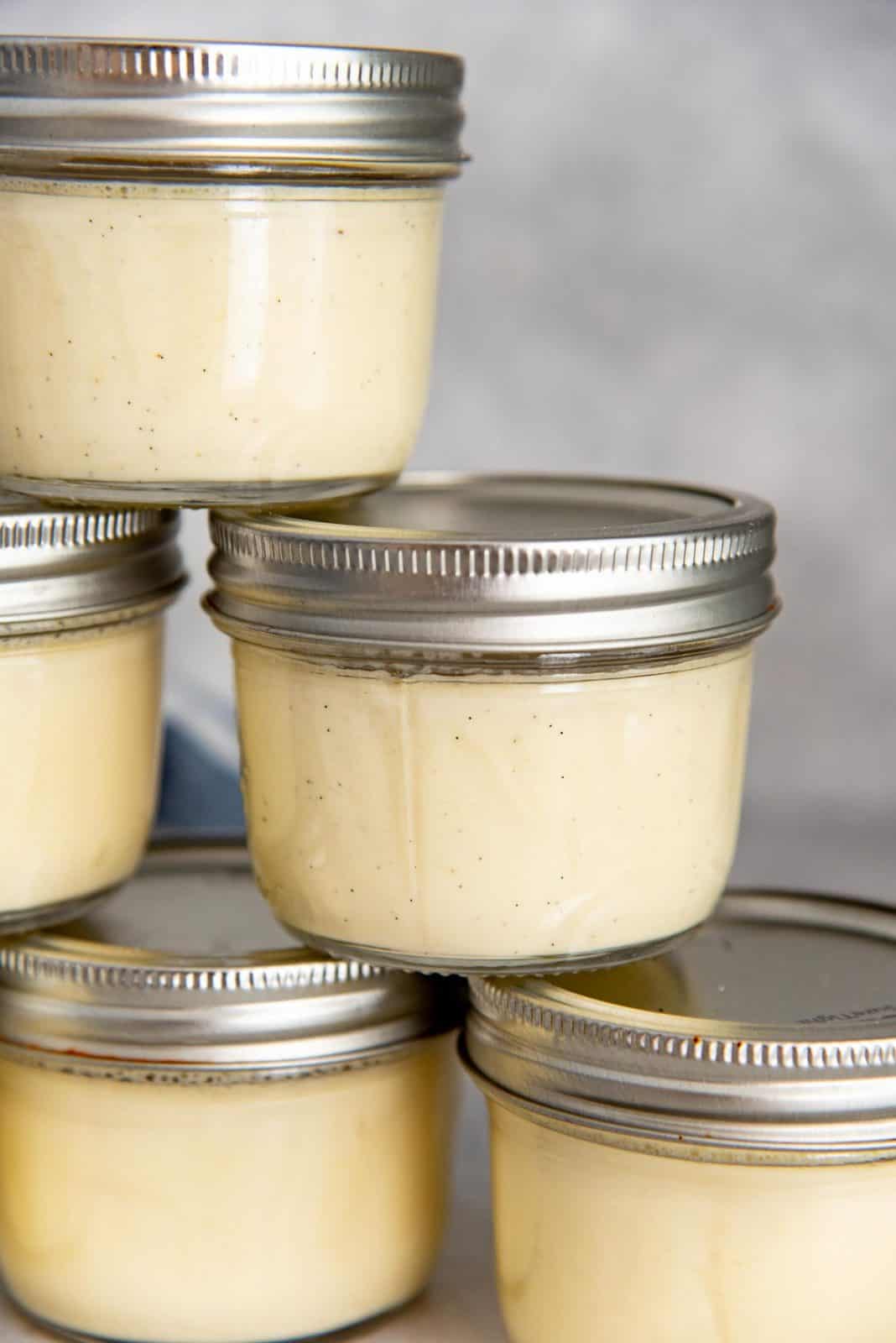 Five small glass jars filled with creamy yellow creme brulee custard, topped with silver lids.