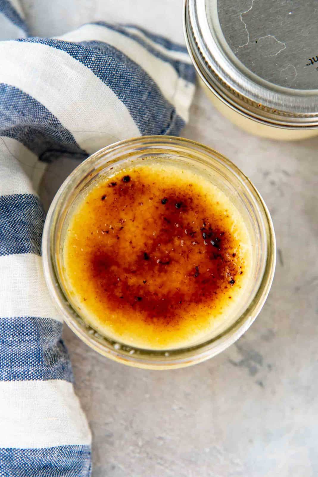 Overhead view of a jar filled with crème brûlée and a caramelized sugar layer top, and a blue and white striped cloth nearby.