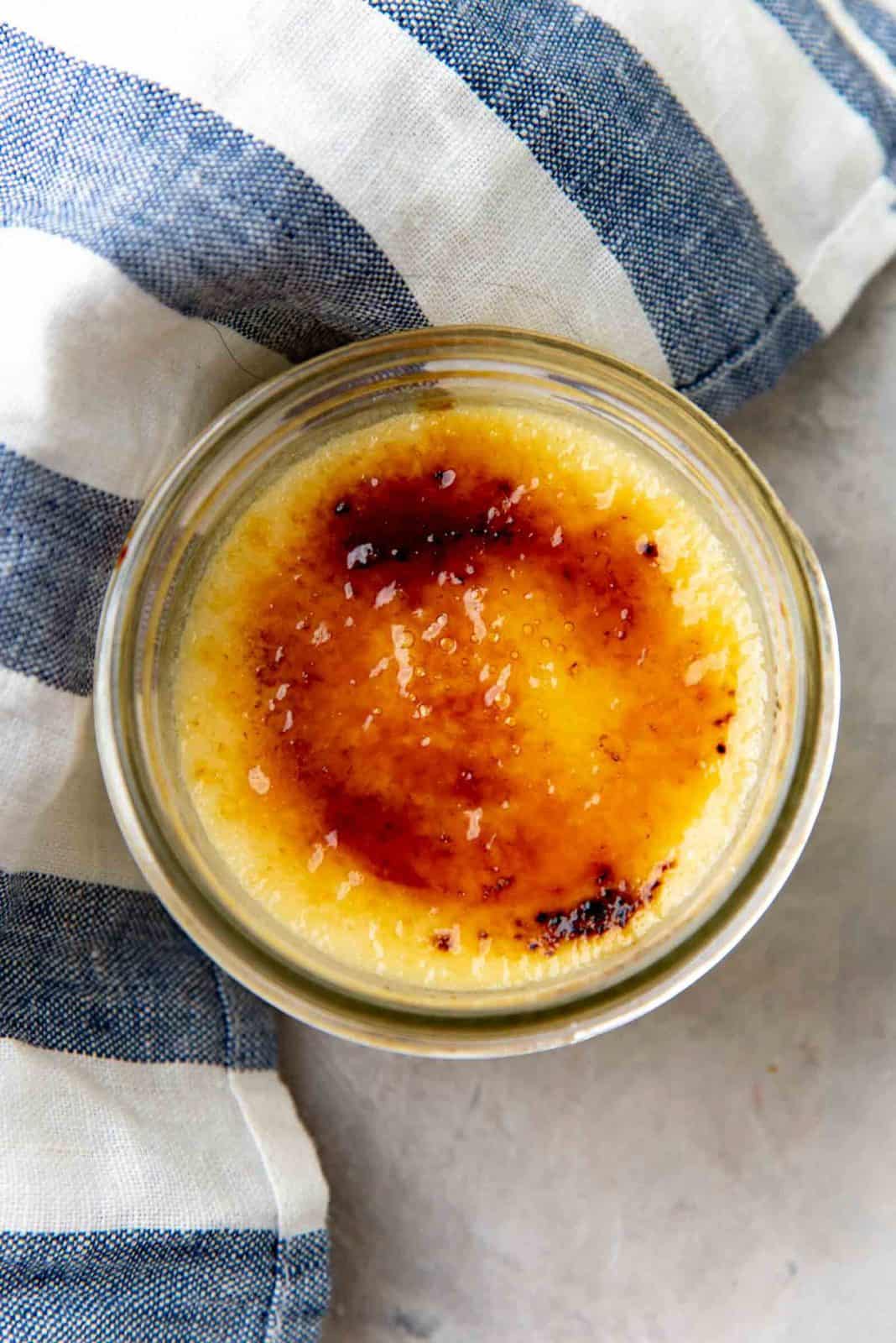 Small glass dish of crème brûlée with a caramelized sugar top, placed on a gray surface next to a blue and white striped cloth.