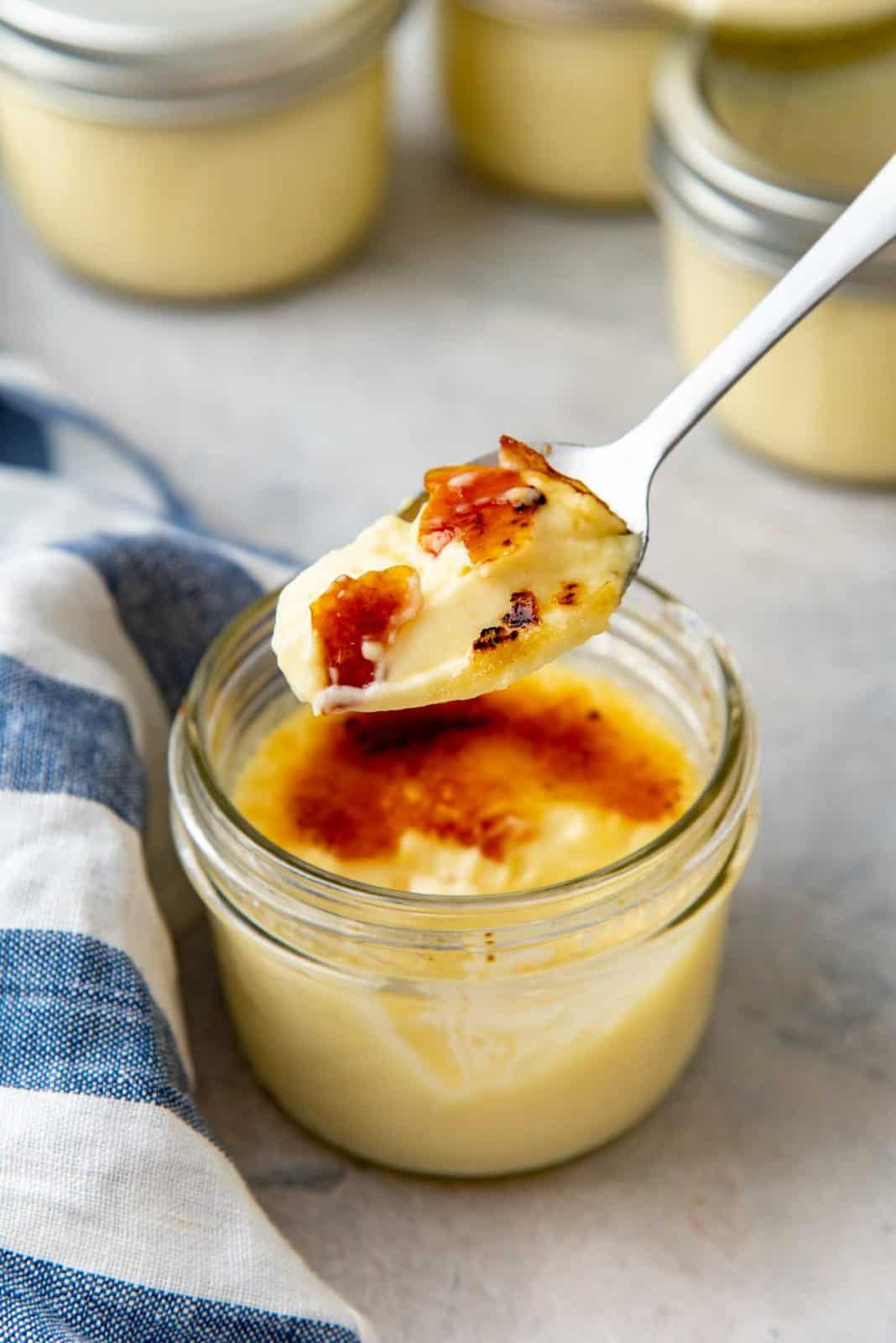 A spoon lifts a creamy crème brûlée with caramelized top from a glass jar, with a striped cloth in the background.