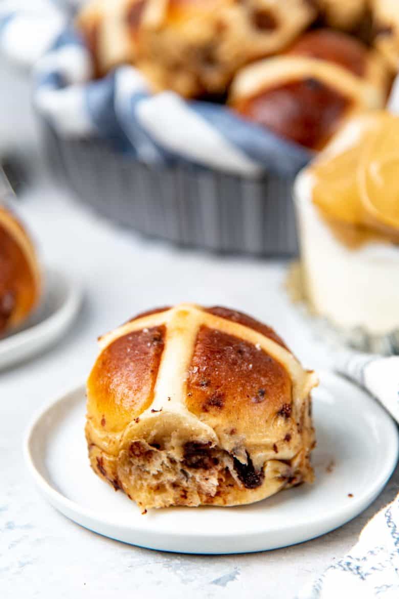 A chocolate chip hot cross bun on a small white plate in the foreground, with coffee and more buns in the background.