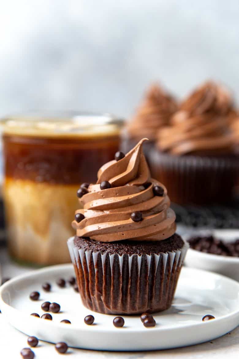 A chocolate cupcake, frosting with chocolate buttercream frosting swirl, with chocolate pearls as decoration on a white plate