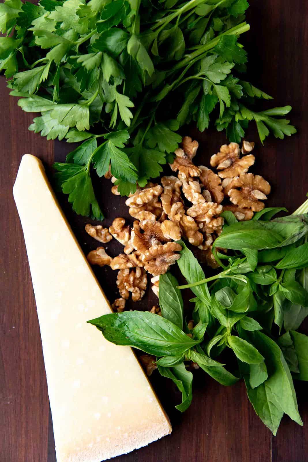 Fresh basil, parsley, walnuts, and a wedge of Parmesan cheese on a wooden surface to make the parsley pesto.