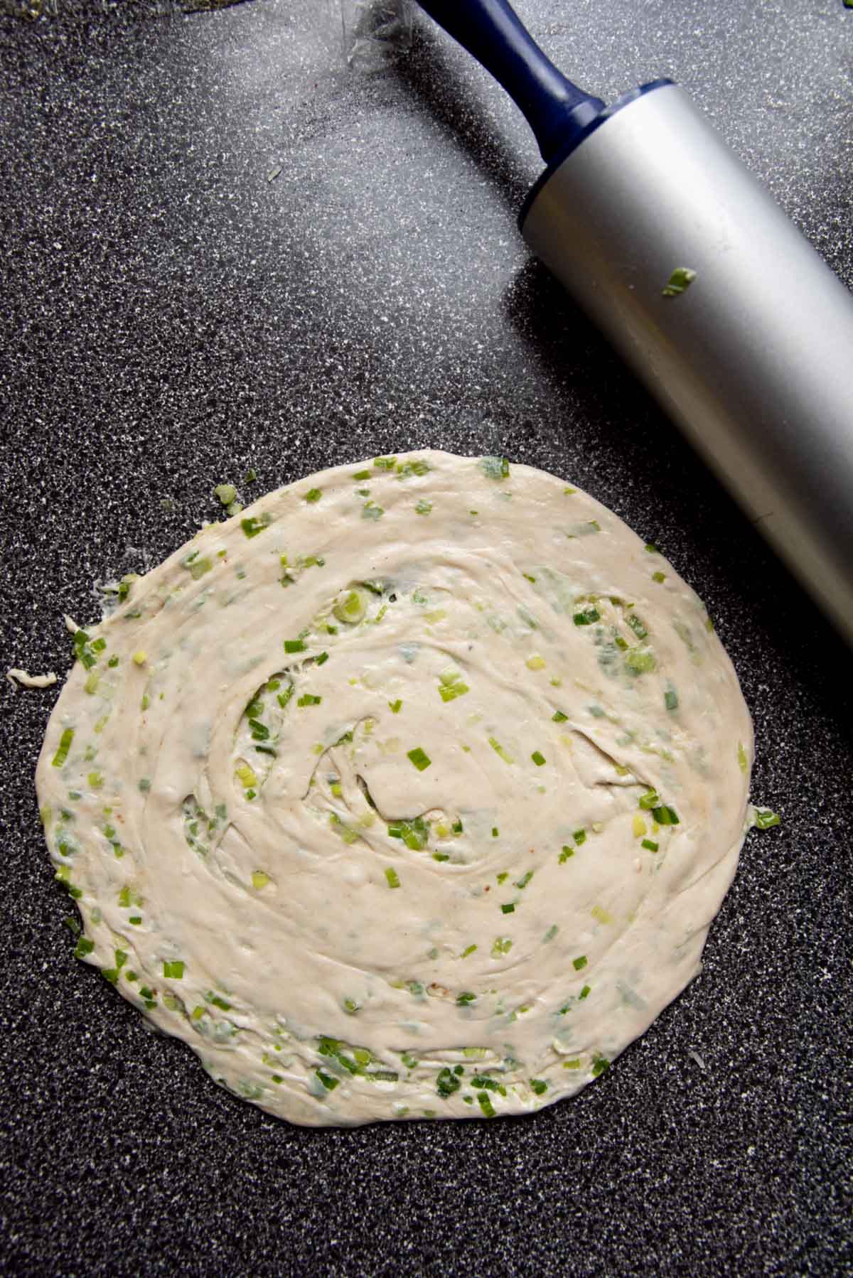 The coiled scallion pancake dough rolled out to a flat bread, with a rolling pin close by.