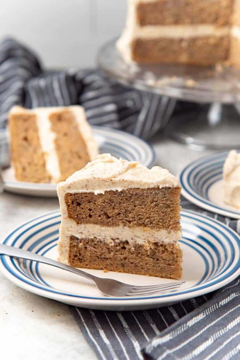 An upright slice of the layered banana cake with brown butter frosting, on a blue rimmed plate with a fork, with another slice in the background.