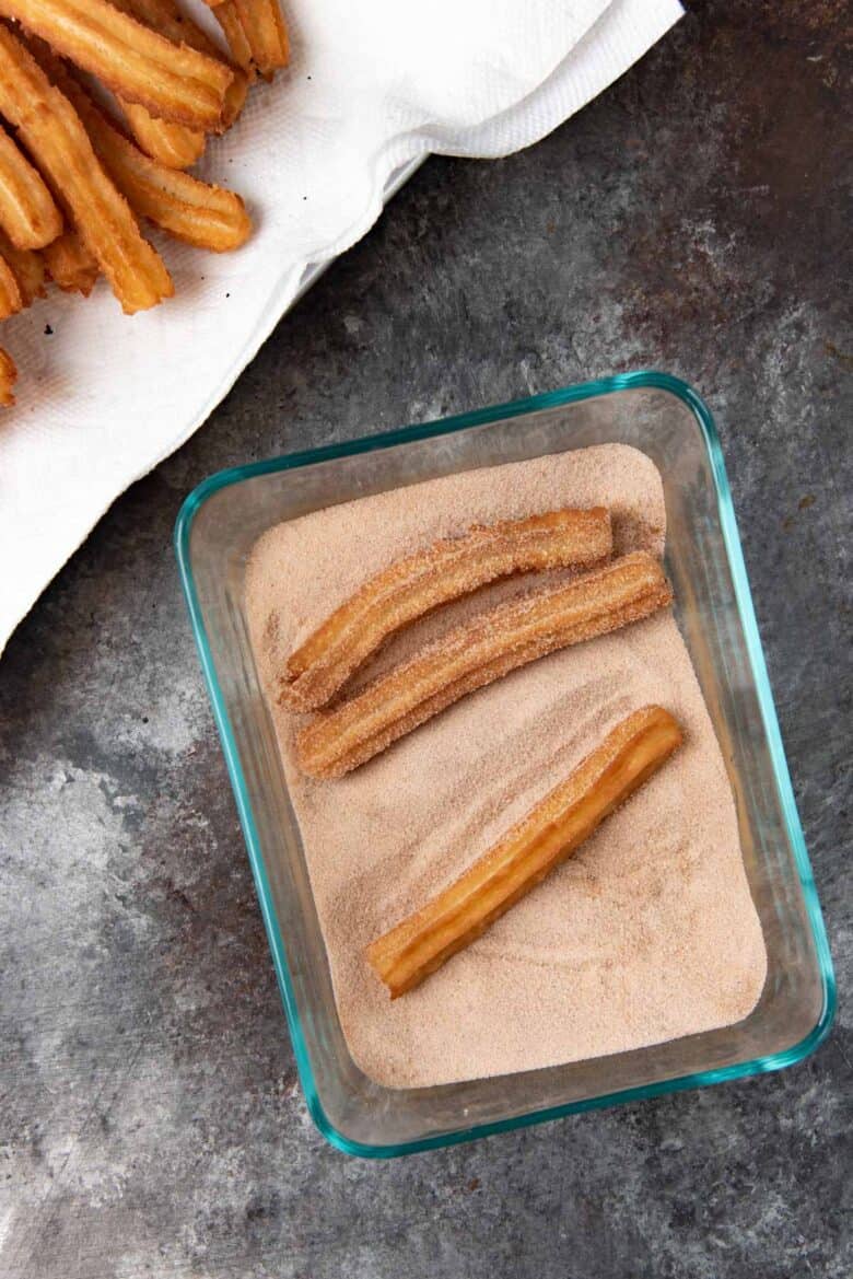 Overhead view of three churros in a cinnamon sugar mix, with more churros stack in another tray next to it.