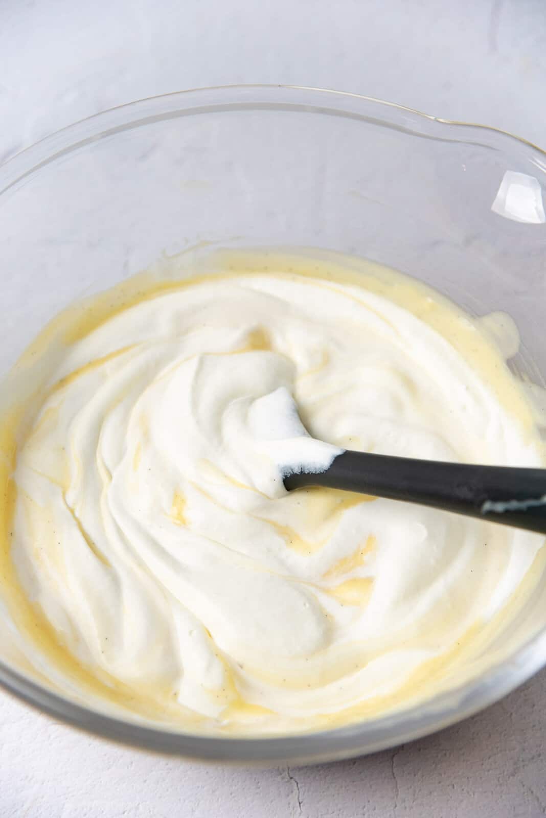 Whipped cream being mixed with the custard base with a dark grey spatula, with white and yellow streaks visible on the surface.