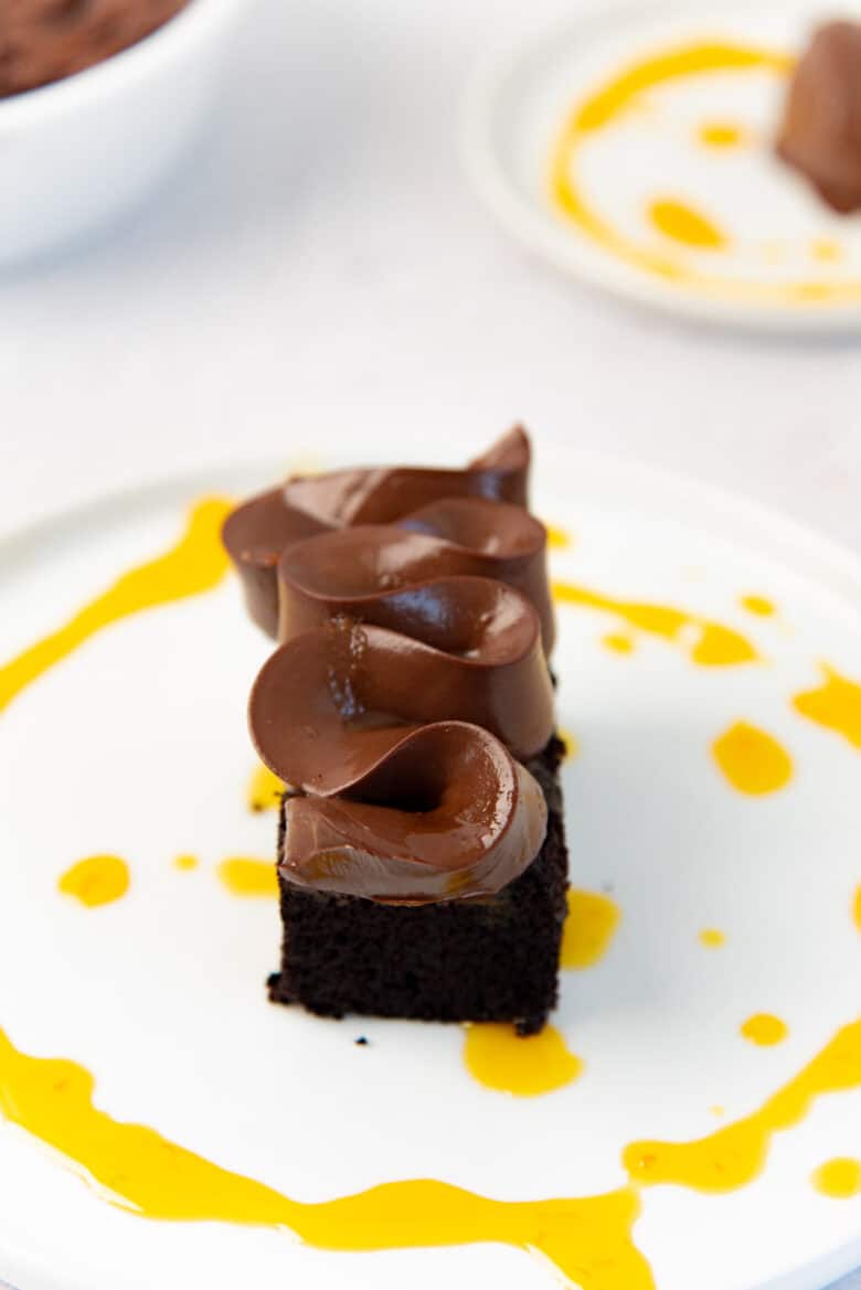 A piece of chocolate cake on a white plate, with chocolate cremeux piped on top of it in a wavy zig zag pattern, with passionfruit syrup in a thin stream around the cake.
