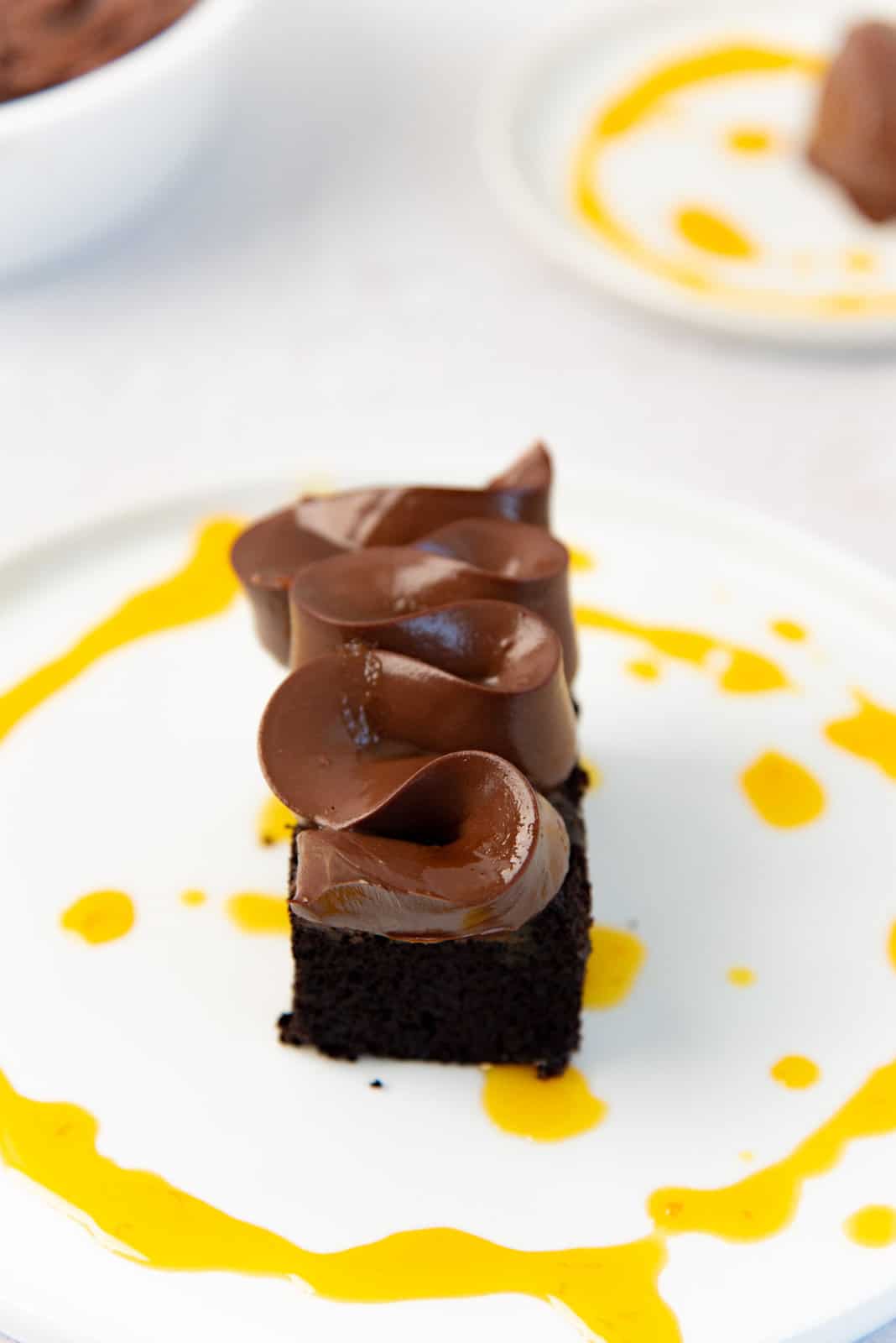 A piece of chocolate cake on a white plate, with chocolate cremeux piped on top of it in a wavy zig zag pattern, with passionfruit syrup in a thin stream around the cake.