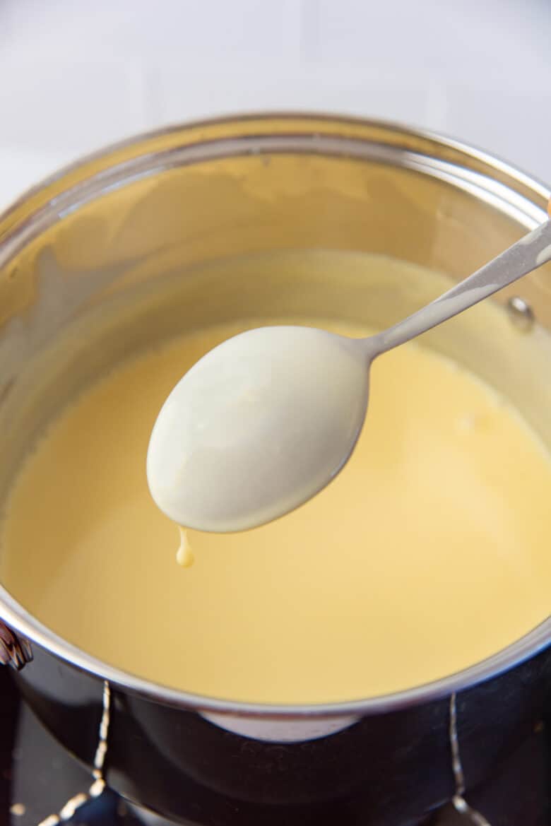 A close up of the back of a large spoon, with a coating of custard on it, with a pot of custard behind it.