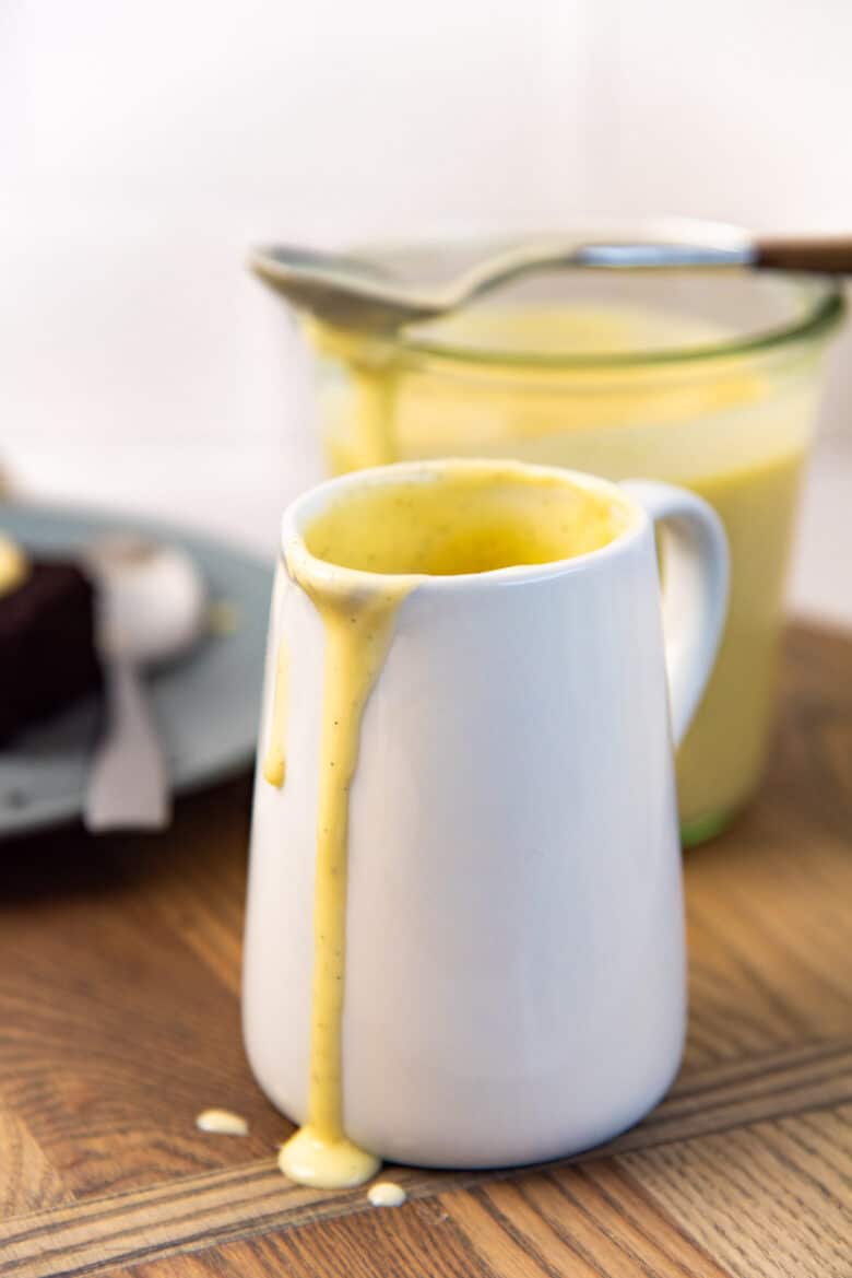 A close view of a white milk jug on a wooden board, that has custard inside, with some custard fallen along the outside near the spout.