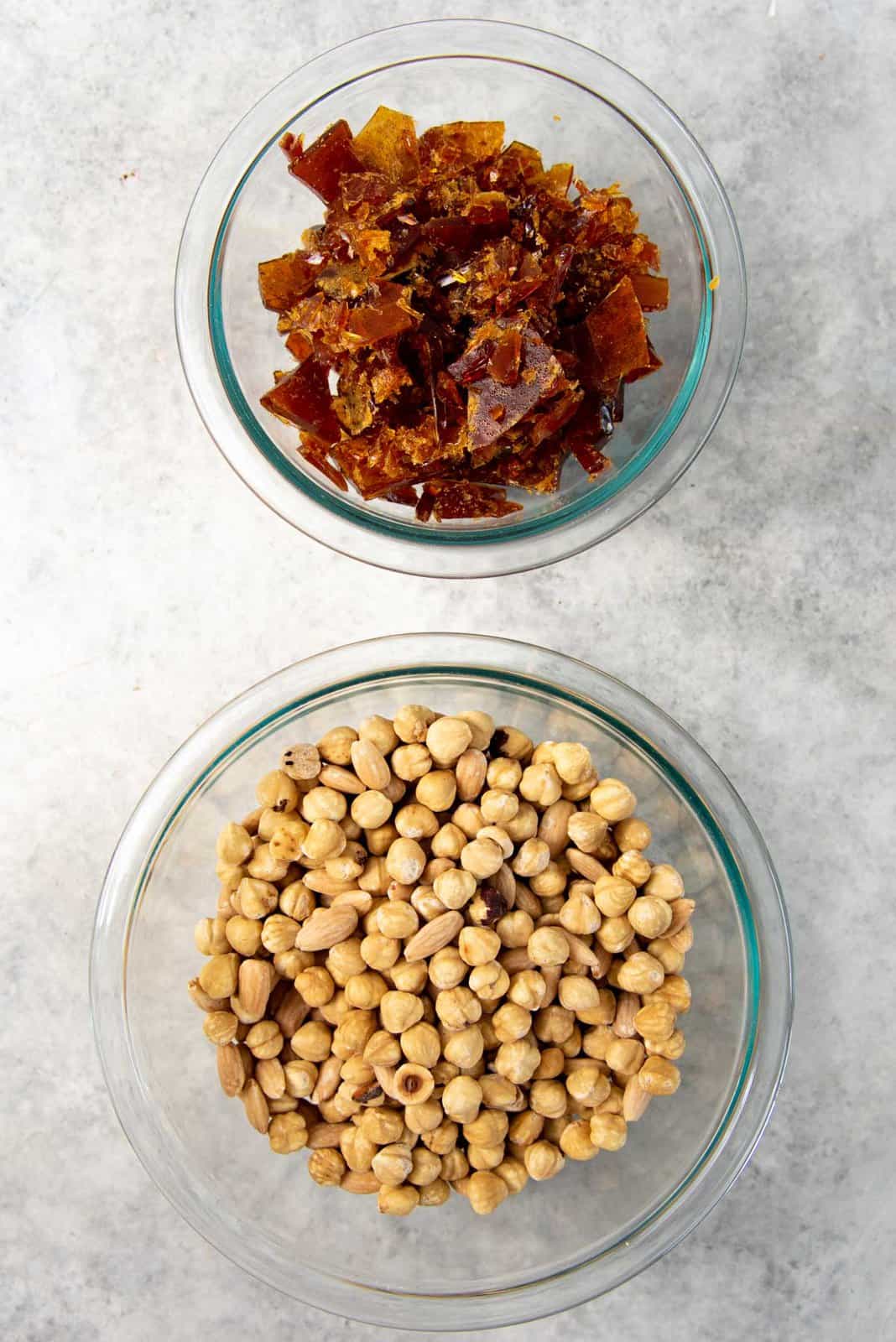 Overhead view of the roasted nuts and the hardened caramel broken into pieces in separate glass bowls.