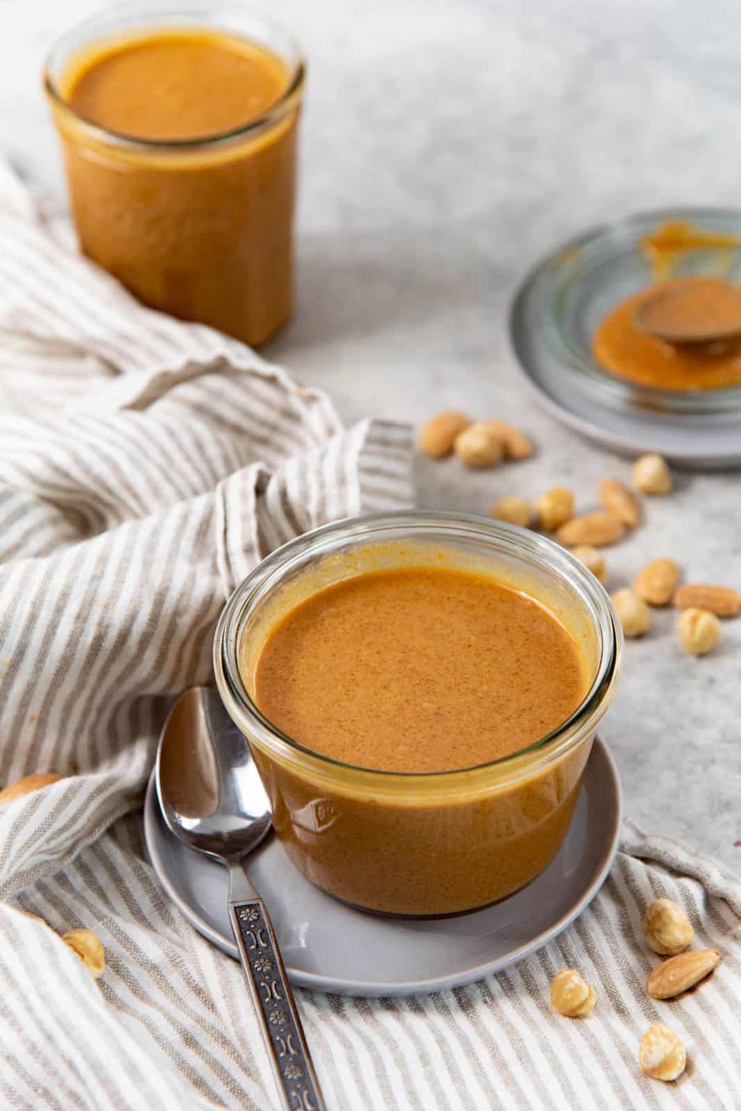 Angled view of the short, small glass jar filled with praline paste placed on a grey plate, with a striped cloth napkin and nuts next to it.