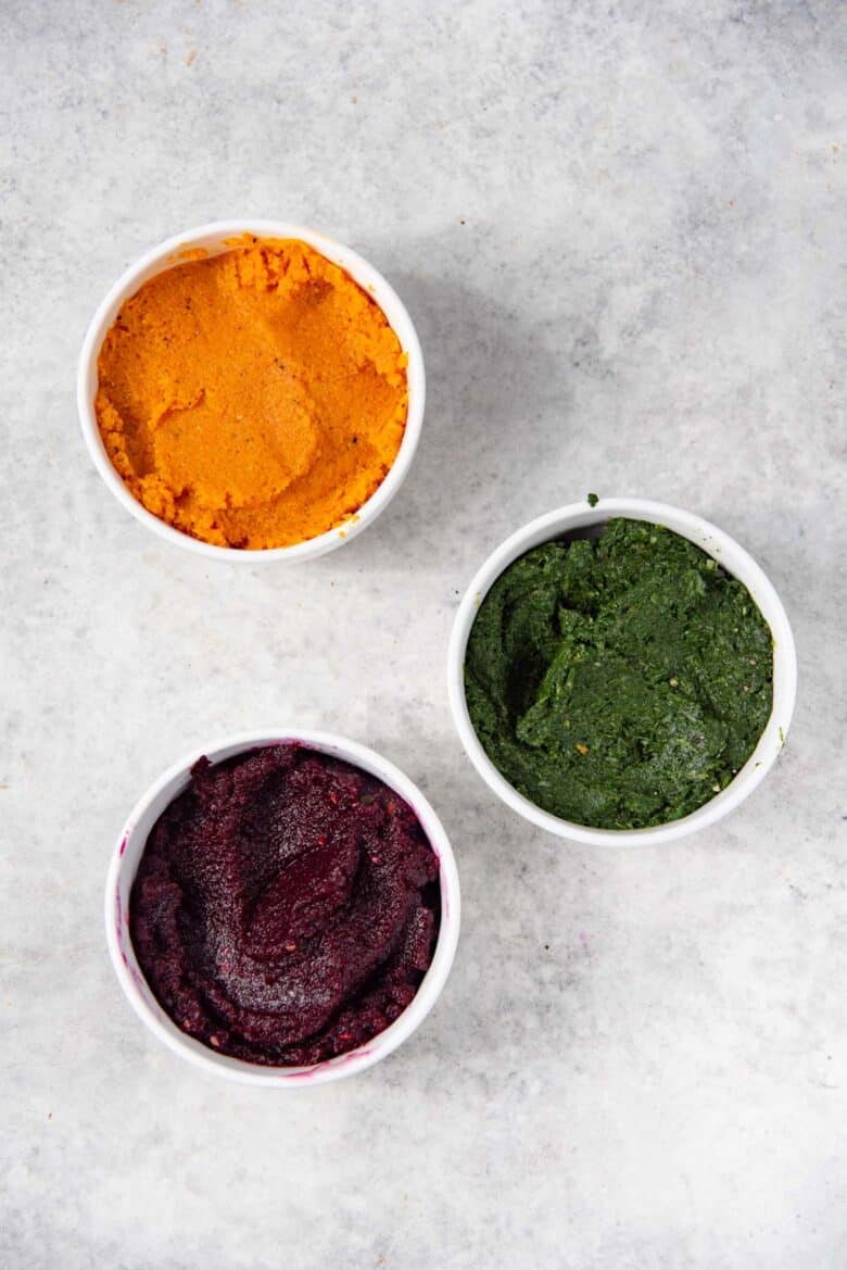 Overhead view of 3 white bowls, with each filled with a carrot paste (top), spinach paste (middle), and beetroot paste (bottom).