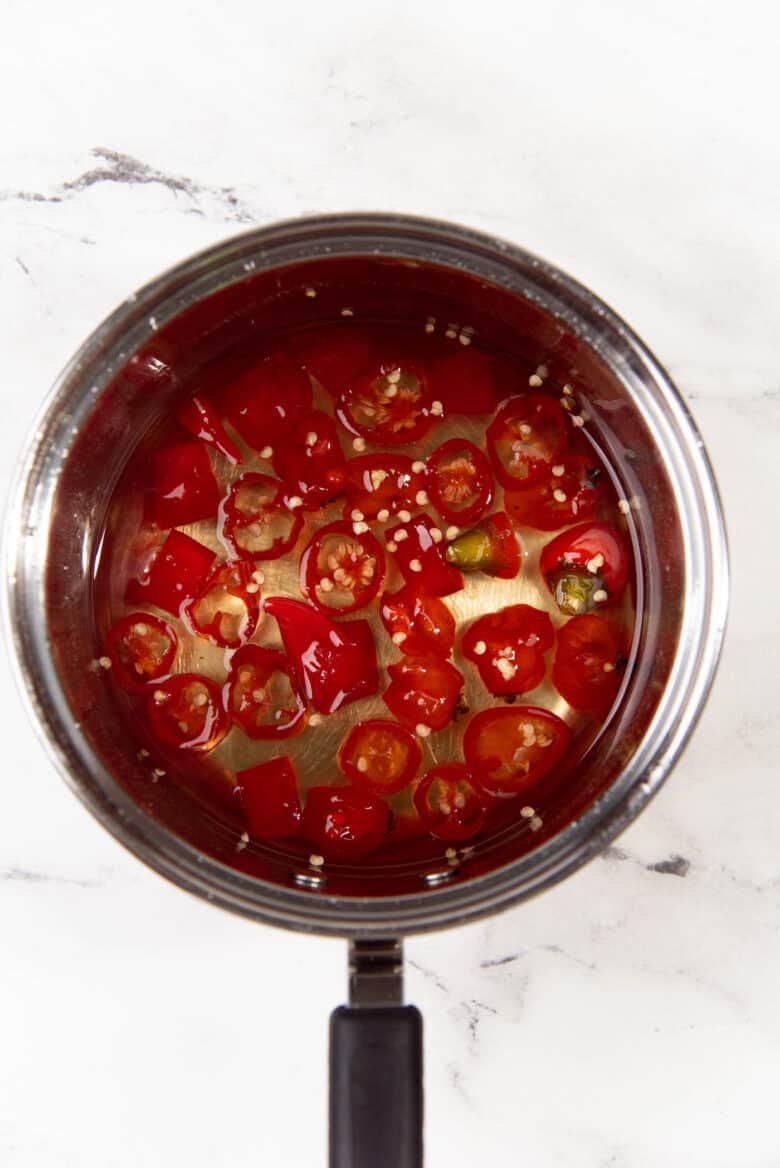 Sliced red chili in a saucepan, floating in a clear liquid that is sugar syrup.