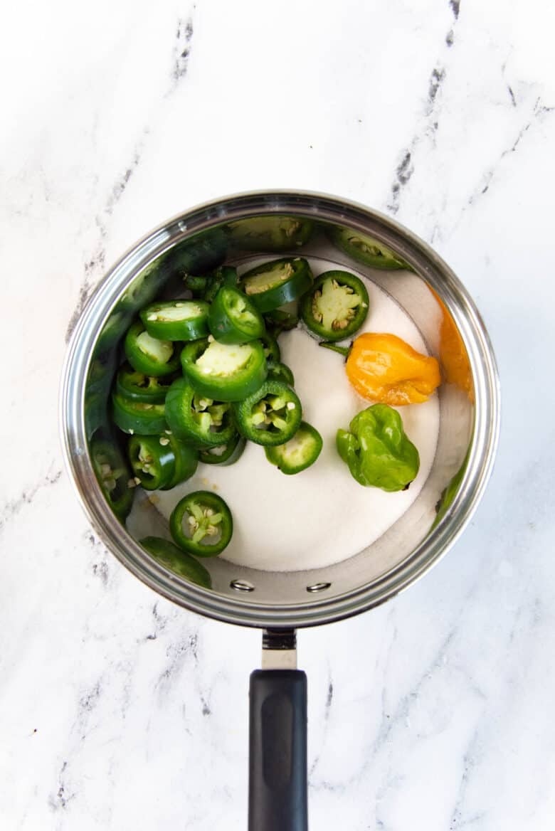 Sliced jalapeno, habanero peppers in a saucepan with white sugar.
