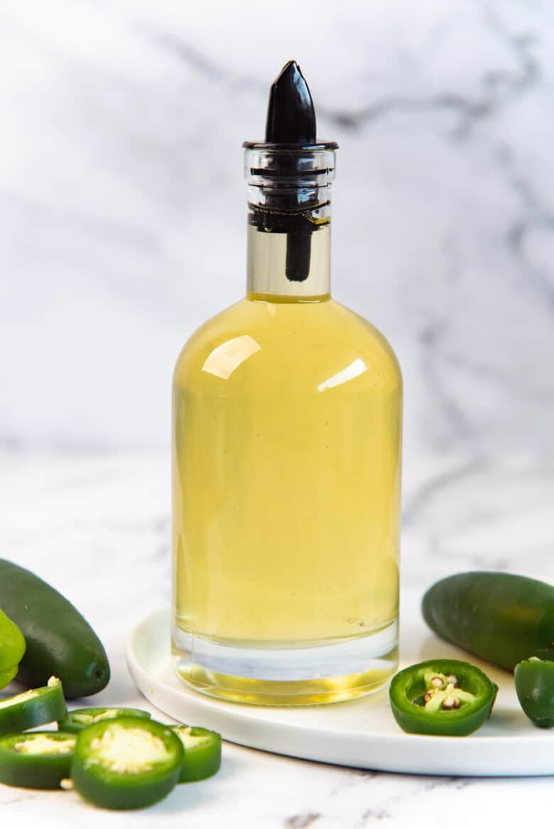 A bottle of Jalapeno simple syrup with a pouring tip, on a white plate, with slices jalapenos in the background and foreground. 