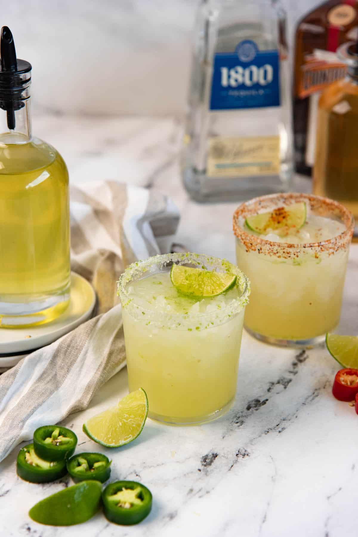 Two glasses of spicy margaritas, made with spicy simple syrup, with slices jalapeno and lime in the foreground.