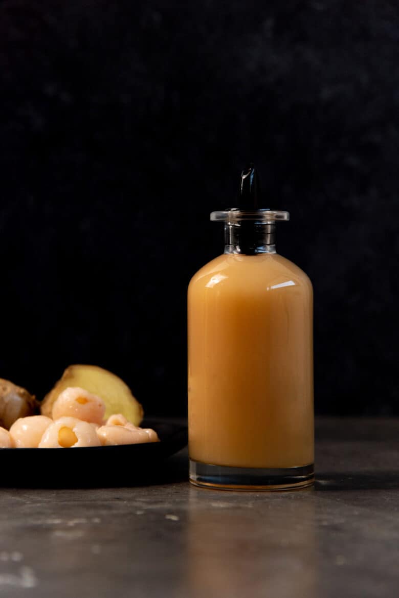 A syrup bottle with a yellowish white syrup on a table top with lychee in a small black saucer next to it.