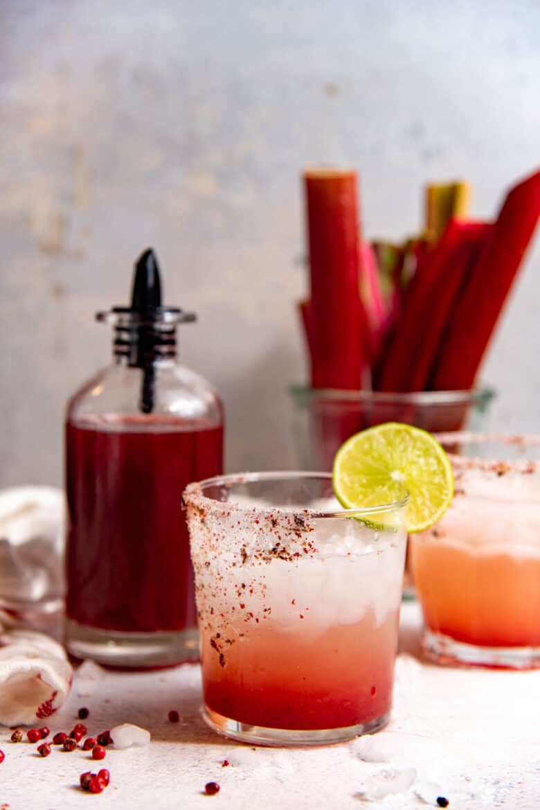 An ombre rhubarb margarita in a glass, with the rhubarb syrup in the background and rhubarb stalks in the background.