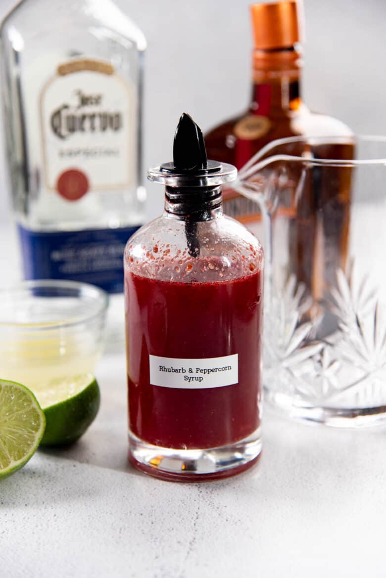 Peppercorn and rhubarb syrup in a bottle with margarita ingredients in the background.