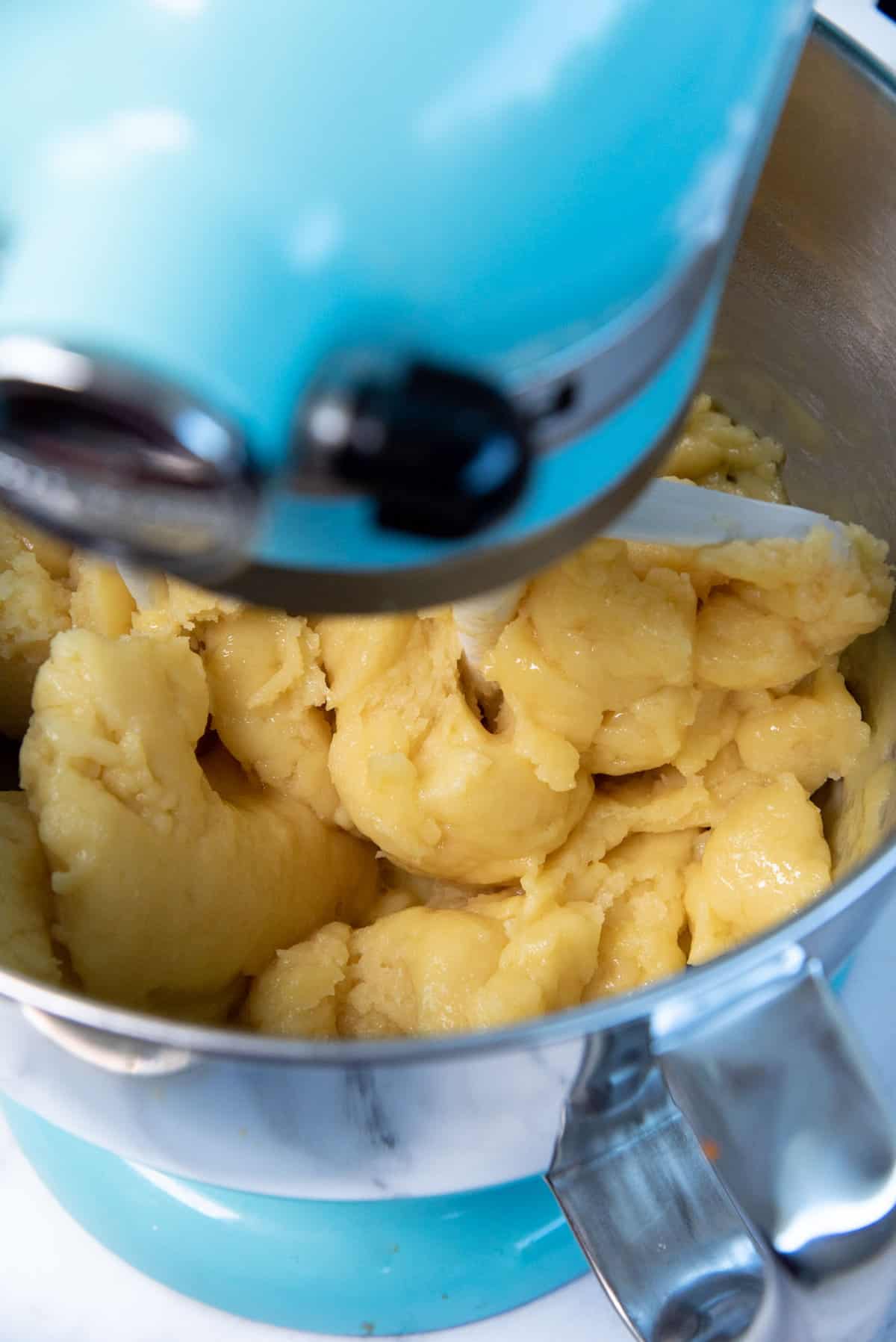 A close up of the choux pastry dough in the bowl of a stand mixer, while eggs are being mixed through.
