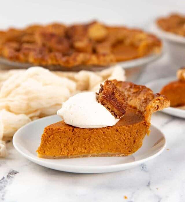 A slice of classic pumpkin pie , served with chantilly whipped cream and sugar crusted pie bites, with the whole pie in the background.