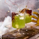 Two green hocus pocus cocktails with spooky smoke and a witch hand prop in the background.