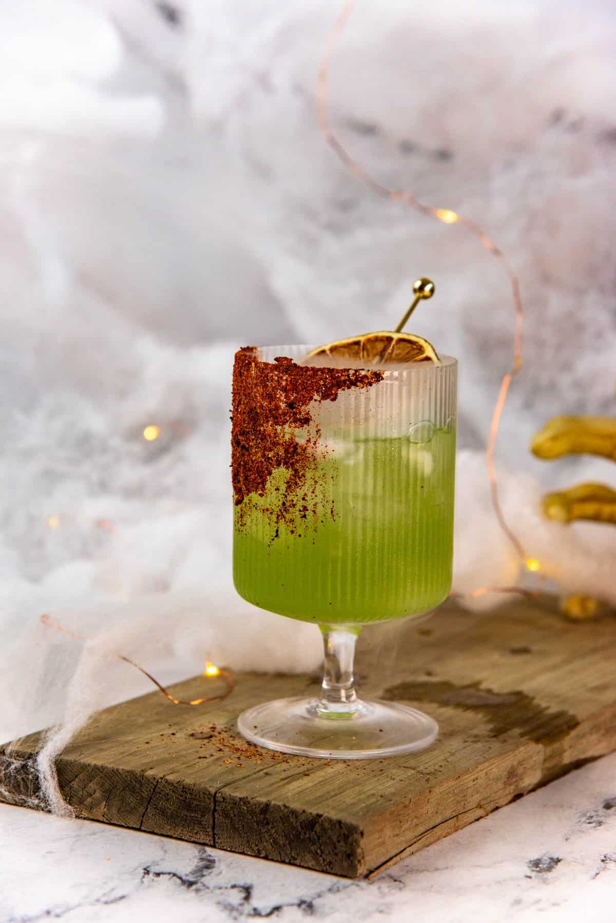 A side view of a green cocktail served in a goblet glass with a chili salt rim, with fairy lights and cobweb fabric in the background, with halloween decorations.