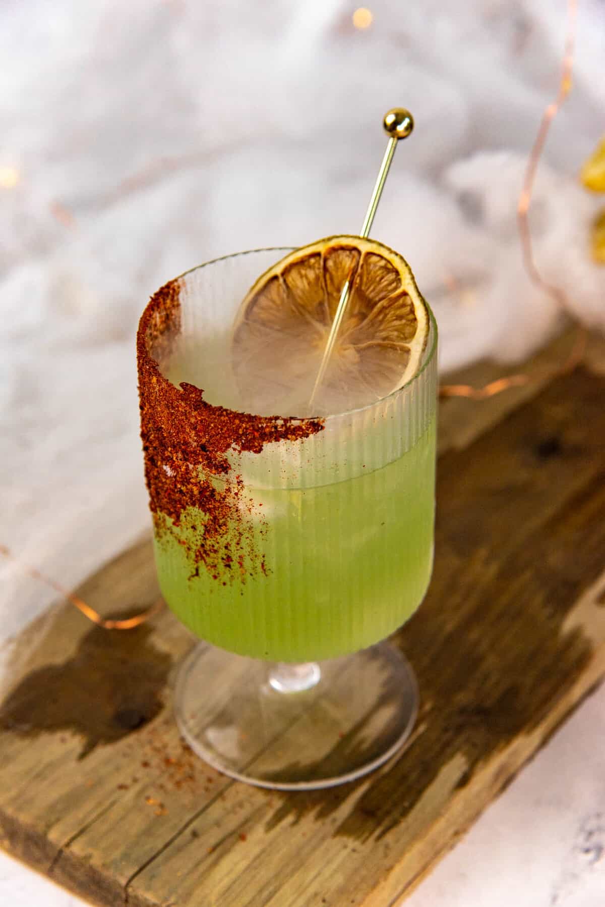 Green colored tequila based halloween cocktail served in a goblet with chili salt rim, served with a stirrer, on a wooden board.