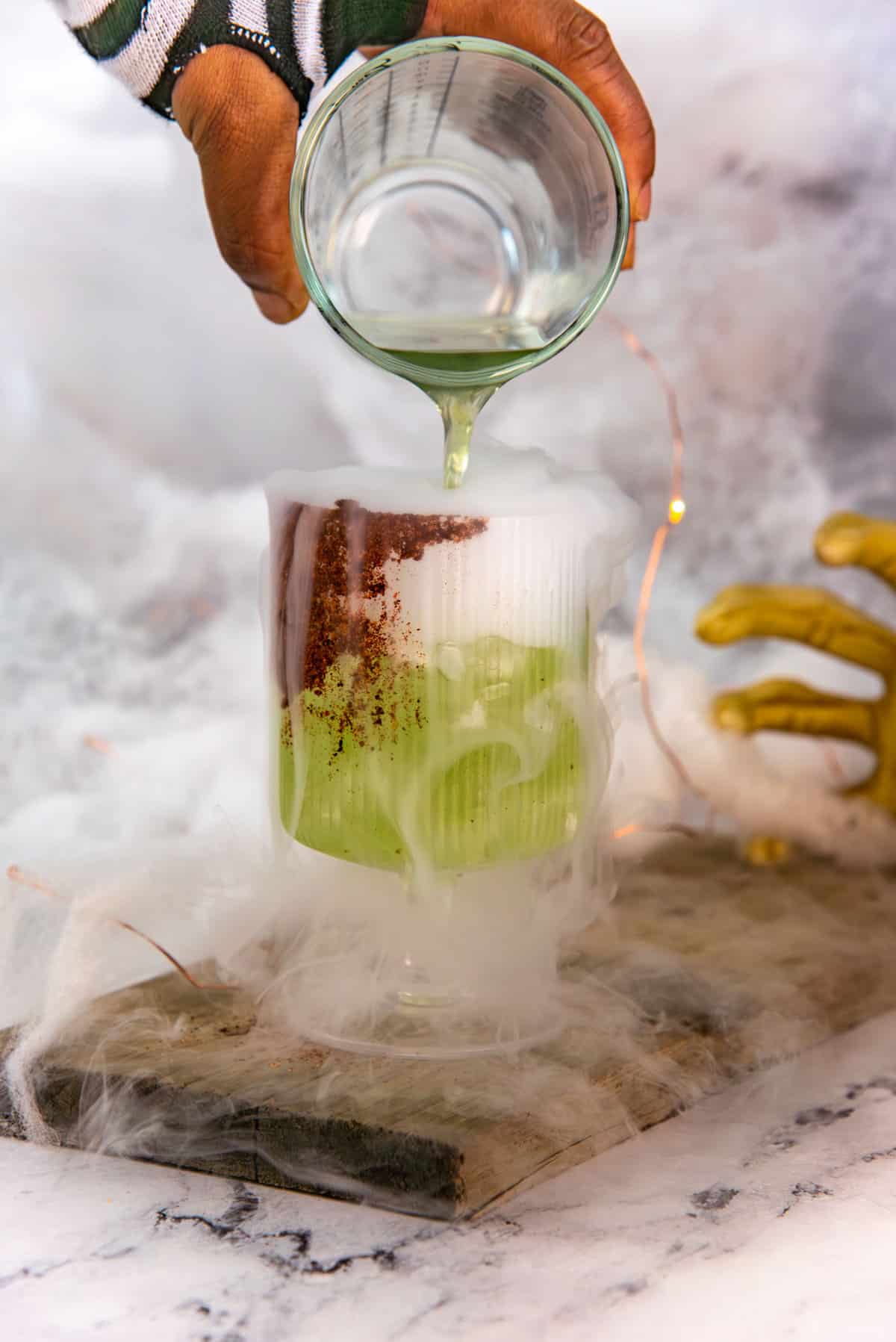 Pouring the green yellowjacket cocktail into a chili salt rimmed goblet glass with smoke spilling out of the glass.