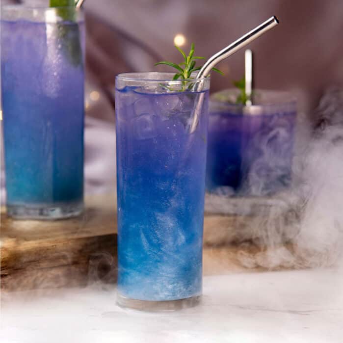 Halloween themed cocktail "Witches Brew" drink, served in a glass with blue to purple color gradient, square image.