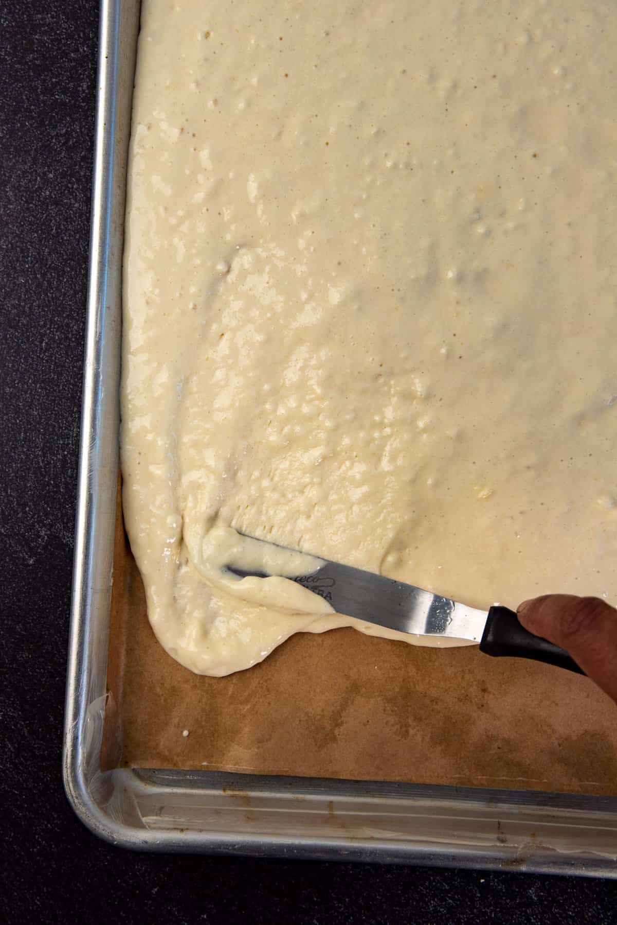 A close up of the pancake batter being spread in the half sheet pan using a small spatula.