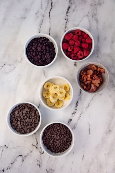 An overhead view of 6 bowls showing different toppings for waffles. Clockwise from top, frozen blueberries, fresh raspberries, cooked bacon, mini chocolate chips, chocolate chips.