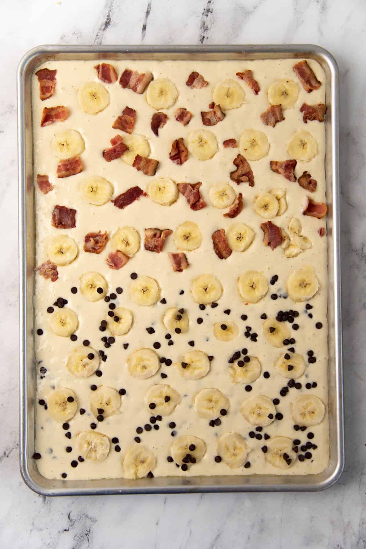 An overhead view of the pancake batter spread evenly on a half sheet pan, with sliced banana and bacon on one half, and banana chocolate chip on the other half.