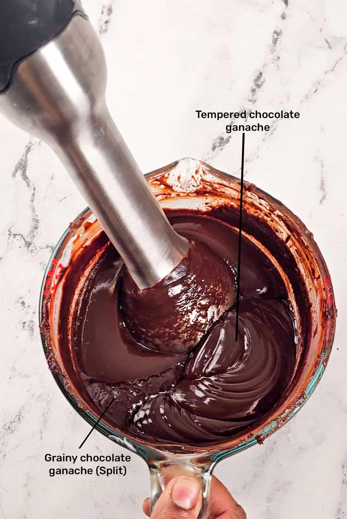 An overhead image of chocolate ganache being mixed with a stick blender to emulsify. The ganache shows two clear textures that are labeled as tempered chocolate ganache and grainy chocolate.