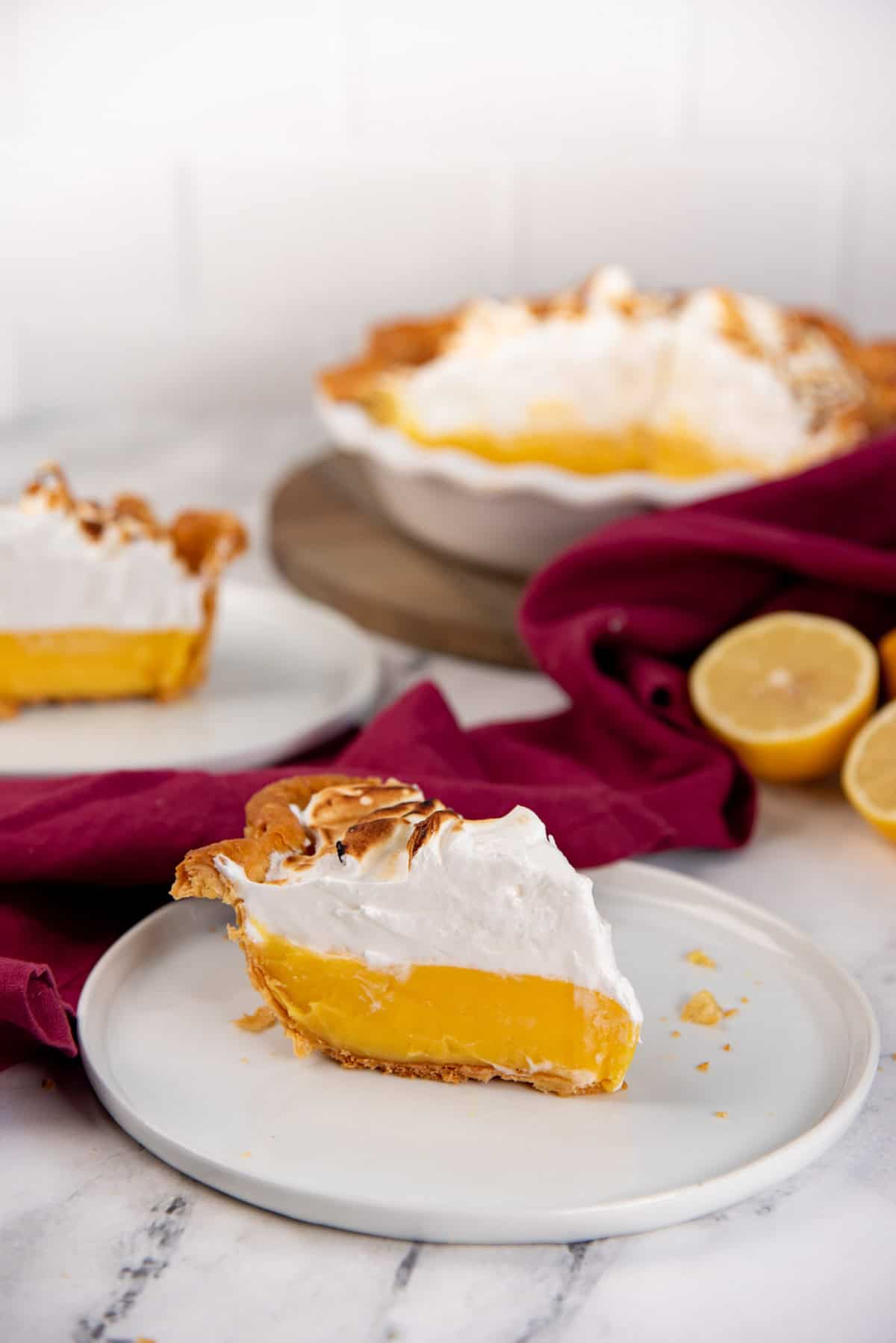 Half of lemon meringue pie and slice in the background, with 1 slice in the foreground on a white plate.