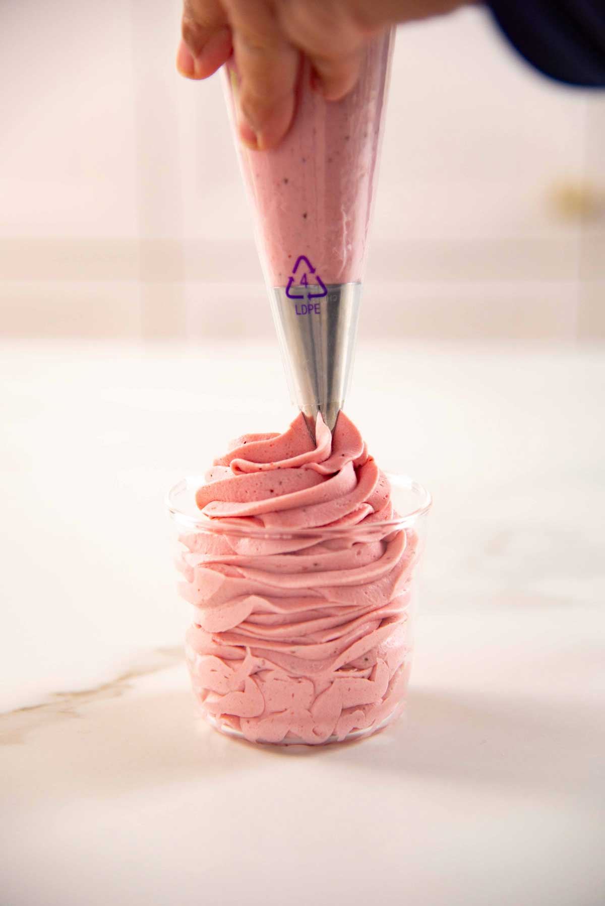 A hand piping a big swirl of pink whipped cream into a glass jar on a white surface.