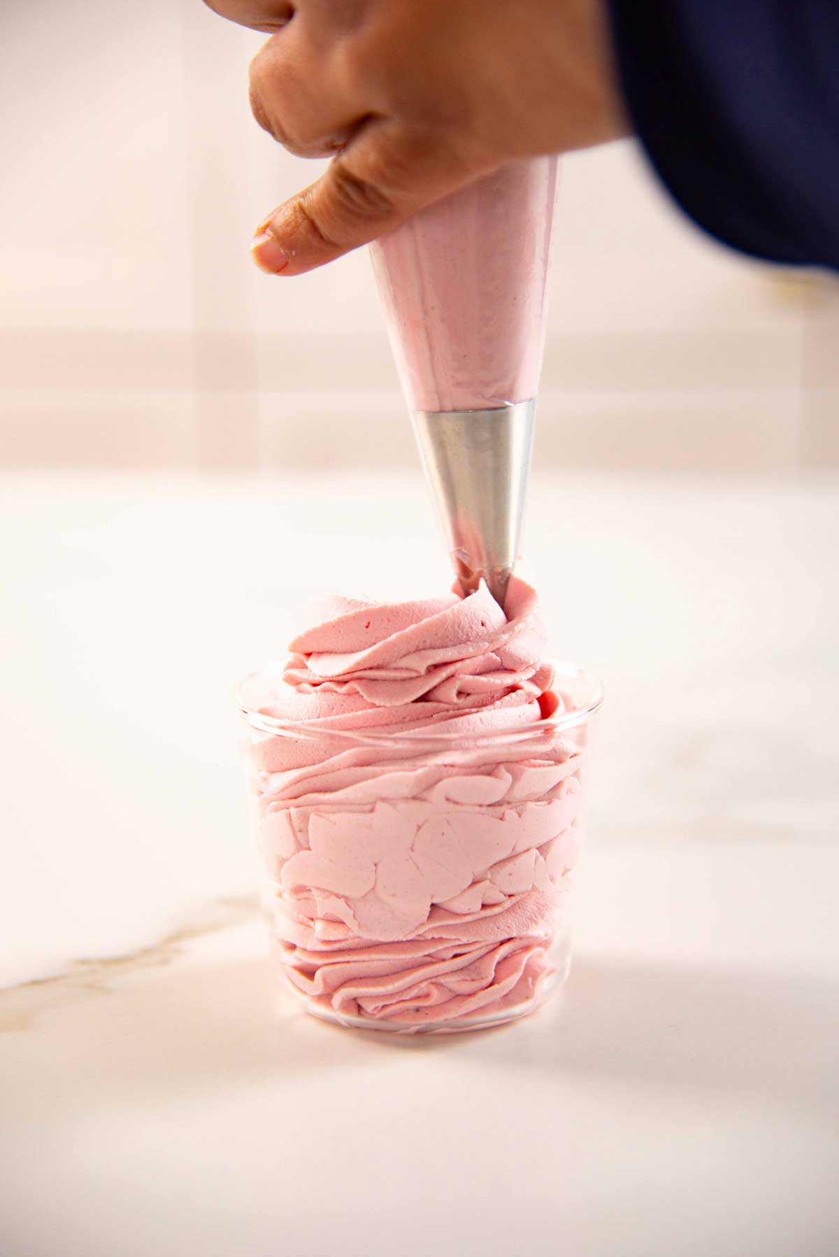 A hand piping a big swirl of pink strawberry whipped cream into a glass jar on a white surface.