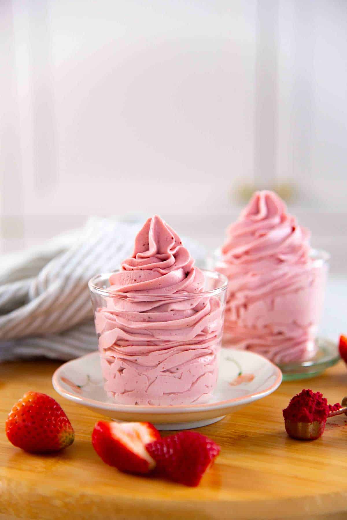 Two glasses filled with swirled pink strawberry chantilly cream on a wooden board with fresh strawberries scattered nearby.