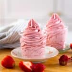 Two glasses filled with swirled strawberry whipped cream on a wooden board with fresh strawberries scattered nearby.