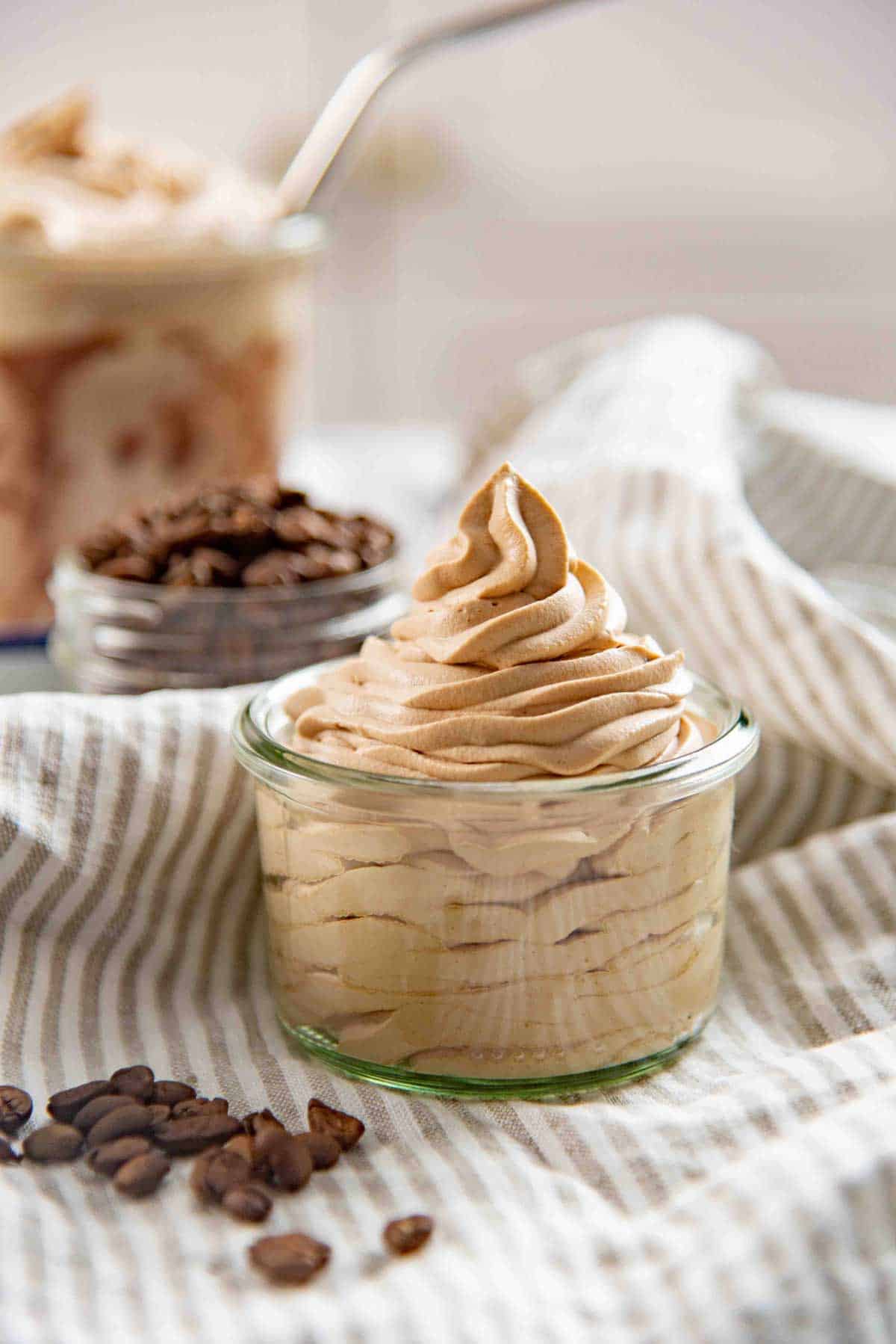 A glass jar filled with espresso whipped cream sitting on a striped cloth, with coffee beans scattered nearby.