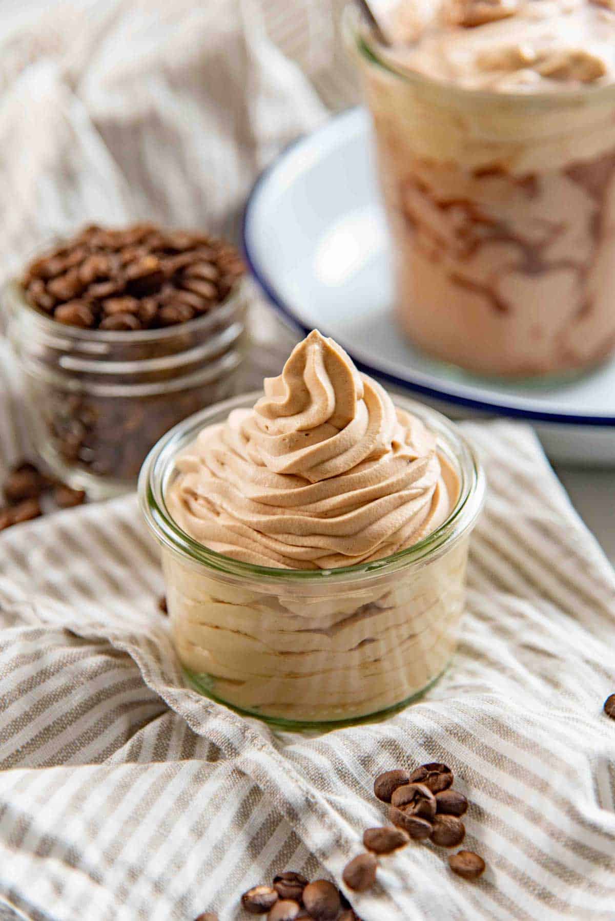 Small glass jar filled with whipped coffee cream on a striped cloth, with coffee beans scattered nearby.