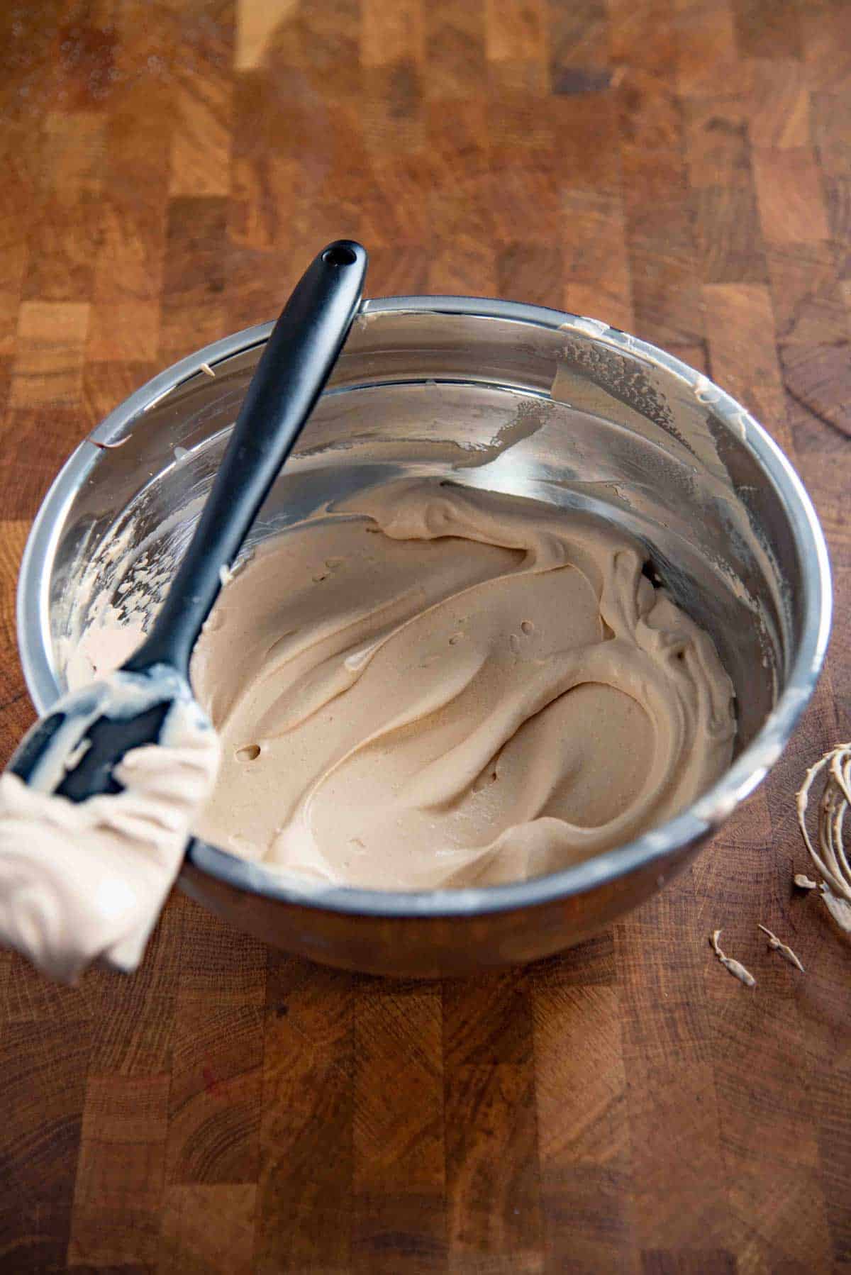 Stainless steel mixing bowl with fluffy coffee whipped cream, and a black rubber spatula resting on top.