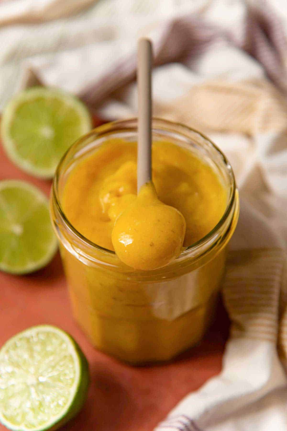 A jar of creamy yellow lime curd with a spoon, surrounded by halved limes and a striped cloth.