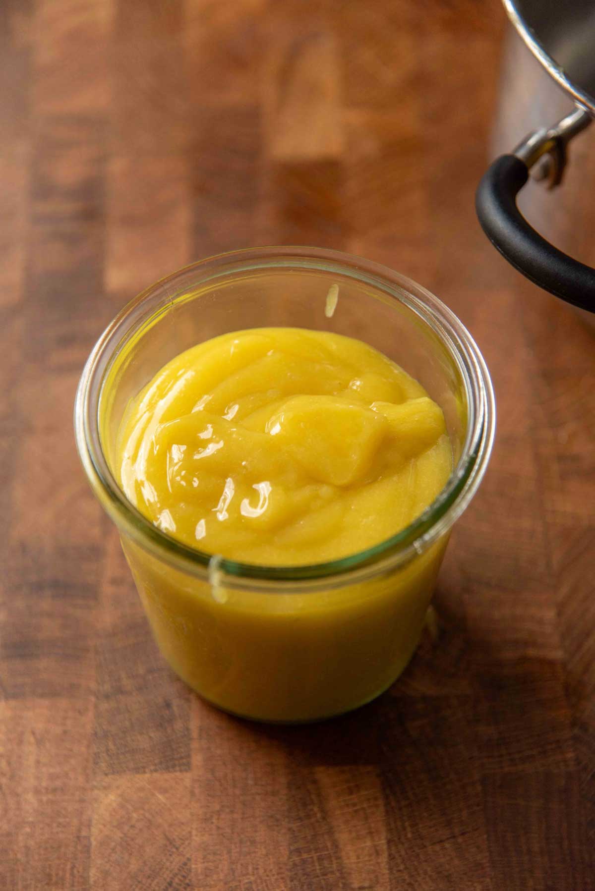 A glass jar filled with smooth, creamy yellow lime curd sits on a wooden counter.