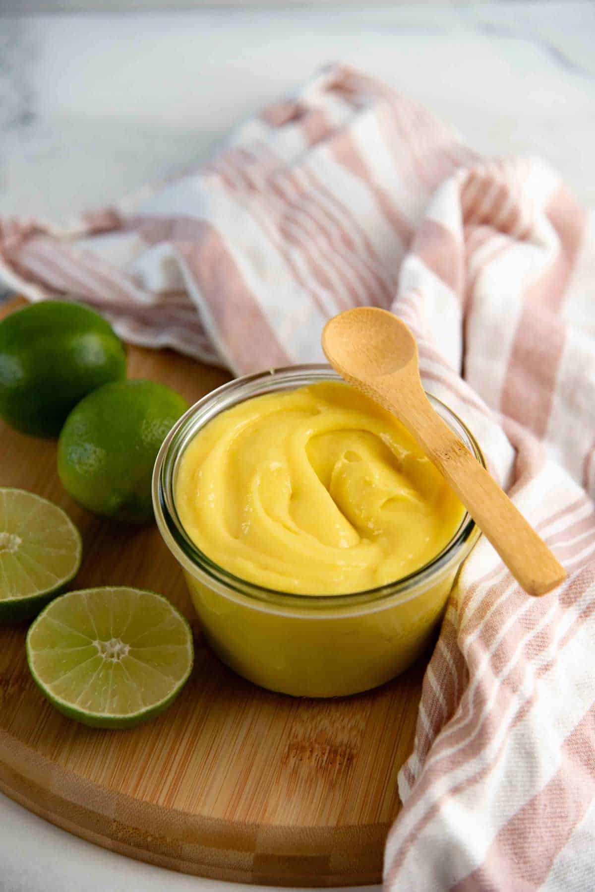 A jar of creamy lime curd with a small wooden spoon on top, placed on a wooden board, with fresh limes nearby.